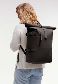 Mochila negra tipo roll-top con correas ajustables, bolsillo frontal y acento de color. Fabricada con material impermeable, con un diseño minimalista.