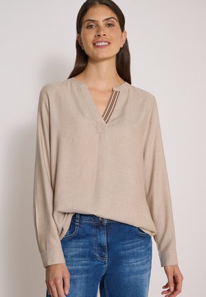 Femme aux longs cheveux foncés et taches de rousseur, portant un chemisier beige à manches longues et un jean bleu, souriant doucement sur un fond uni.
