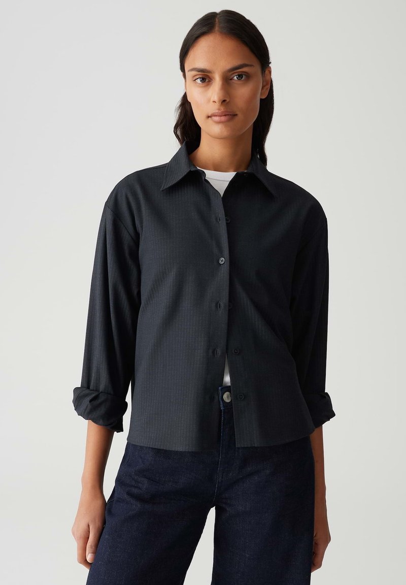 Donna con capelli scuri che indossa una camicia nera con bottoni sopra una maglietta bianca e jeans blu scuri, in piedi contro uno sfondo chiaro e semplice.