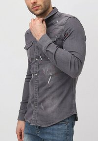 Chemise en denim grise à manches longues, ouverture boutonnée devant, deux poches poitrine, détails usés et éclaboussures de peinture rouges et blanches.