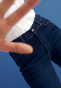 Vaqueros azul oscuro con un diseño ajustado, costuras doradas, un cierre con botón y cremallera visible, y bolsillos frontales.