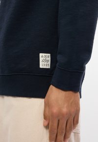 Ναυτικό μπλε φούτερ με ραμμένα μανίκια, διαθέτει υφή υφάσματος και έναν τετράγωνο λευκό ετικέτα με την ένδειξη "MS 1985" στο κάτω αριστερό μέρος.