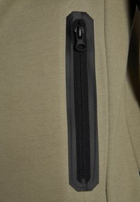 Tissu vert olive avec une poche zippée noire. La fermeture éclair est dotée d'un tirant noir et d'un détail en textile, le tout sur une surface lisse et texturée.