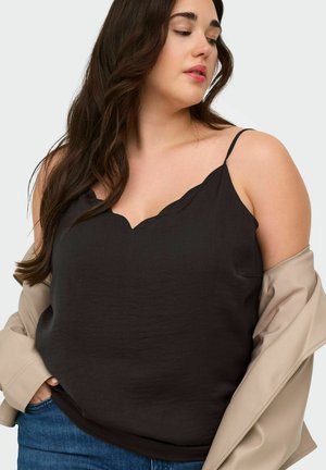 Femme aux longs cheveux foncés portant un caraco noir à bords festonnés, un jean bleu et une veste beige tombée sur les épaules.