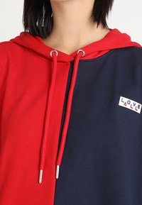 Sudadera bicolor en rojo y azul marino con cordón ajustable, detalles metálicos plateados y un pequeño logo de "LOVE" en el pecho, fabricada en suave tela de algodón.
