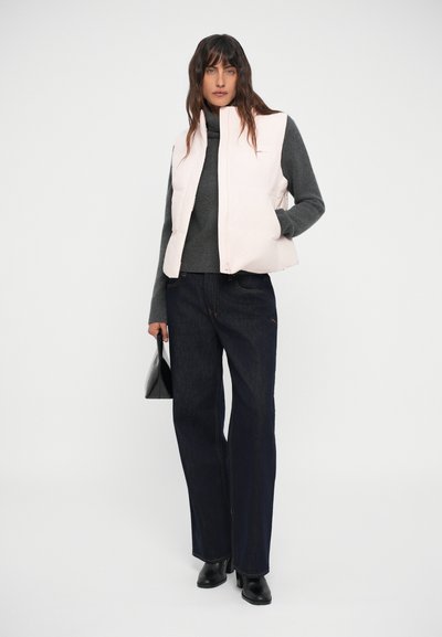 Gilet imbottito rosa chiaro sopra un maglione grigio scuro, abbinato a jeans a gamba larga blu scuro e stivali neri alla caviglia. Borsa nera come accentuazione.