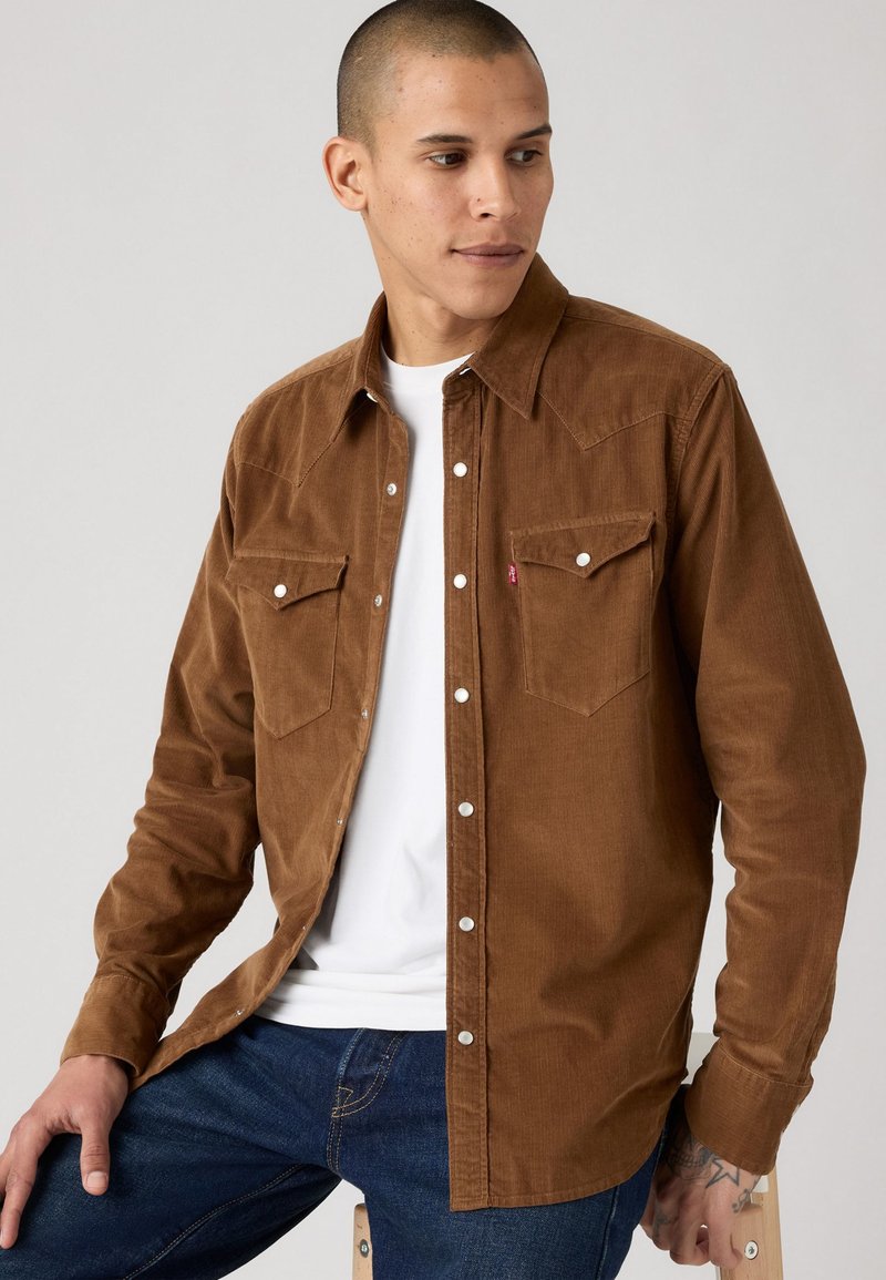 Man in een bruine corduroy button-up shirt over een wit T-shirt en blauwe jeans, zittend op een houten kruk, kijkend naar zijn linkerzijde.