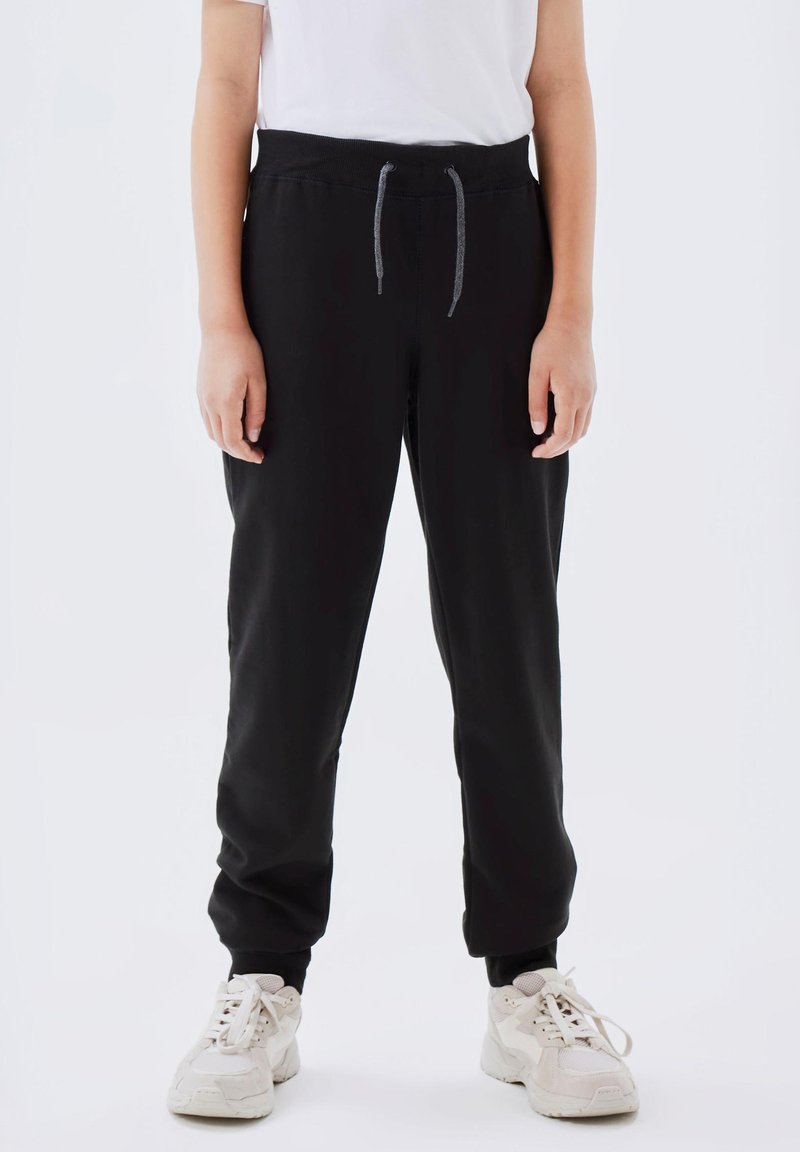 Schwarze Jogginghose aus weichem Stoff, mit einem elastischen Bund und einem Kordelzug sowie gerippten Bündchen, kombiniert mit weißen Sneakers.