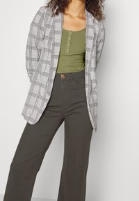 Černobílý kostkovaný blejzr s dlouhými rukávy, nošený přes zelený žebrovaný top. V páru s kalhotami s vysokým pasem v tmavě olivově zelené barvě.