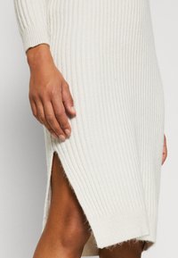 Main reposant sur une robe blanche en maille côtelée avec une fente latérale, montrant un teint de peau foncé et un détail de tissu texturé.