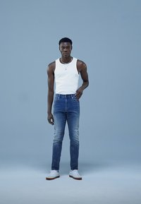 Fehér ujjatlan felső, kék, elszíneződött, szűk farmernadrág és fehér tornacipő barna talppal. A modell világoskék háttér előtt áll.