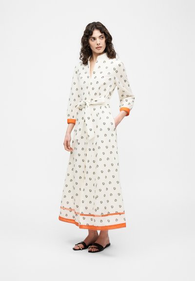 Femme portant une longue robe blanche avec de petits motifs floraux, bordure et poignets orange, taille ceinturée, debout et regardant sur le côté.