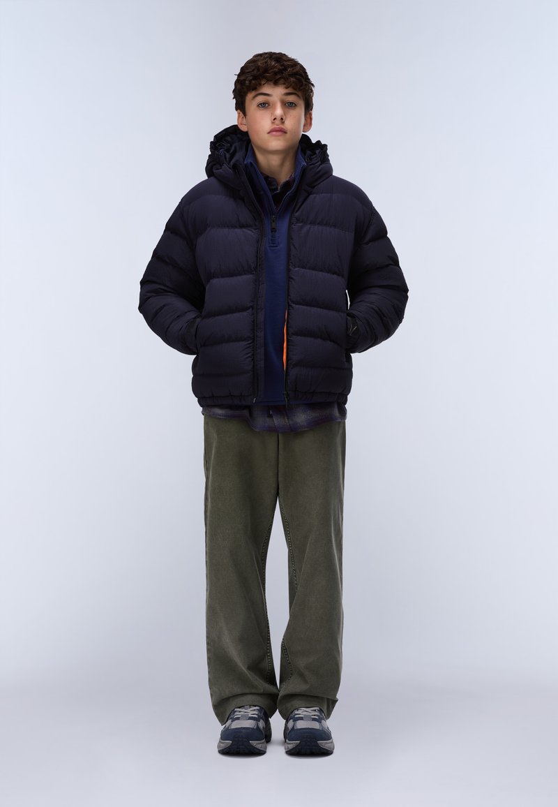 Junge steht mit den Händen in den Taschen, trägt eine schwarze Daunenjacke, ein blaues Hemd, grüne Hosen und Turnschuhe vor einem einfärbigen Hintergrund.
