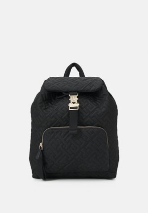 Sac à dos matelassé noir avec poche zippée à l'avant et fermeture à boucle dorée, présenté sur un fond gris clair uni.