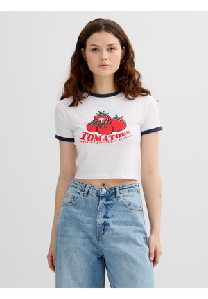 Joven con cabello rizado que lleva una camiseta corta blanca con estampado de tomates y unos vaqueros azules de talle alto, de pie frente a un fondo liso.