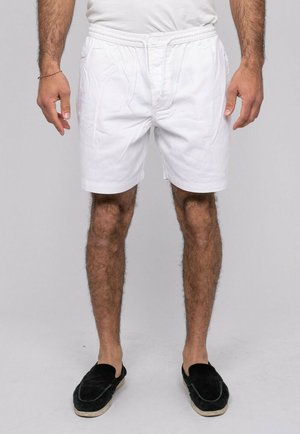 Homme portant un short blanc à taille élastique, un t-shirt blanc uni, des chaussures noires sans lacets, debout devant un fond neutre.