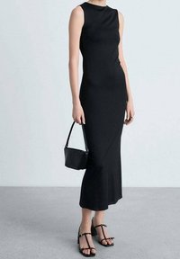 Robe noire sans manches avec une silhouette ajustée atteignant le mollet. Associée à des sandales noires à lanières et à un petit sac à main noir.