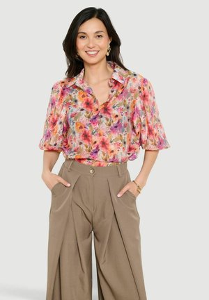 Usmievavá žena s dlhými tmavými vlasmi má na sebe kvetovanú blúzku s krátkym rukávom a béžové nohavice s vysokým pásom a širokými nohavicami, ruky vreckách.