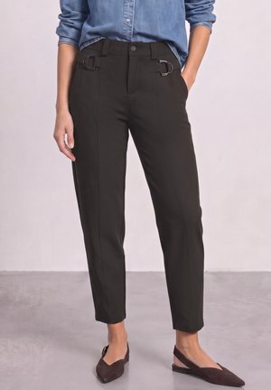 Femme portant un pantalon noir fuselé avec des boucles latérales, coupé aux chevilles, associé à des chaussures slingback à bout pointu foncées et une chemise bleue.