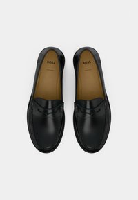 Czarne skórzane mokasyny wsuwane z detalicznym przeszyciem, beżowe wkładki z logo "BOSS", widoczne z góry na białym tle.