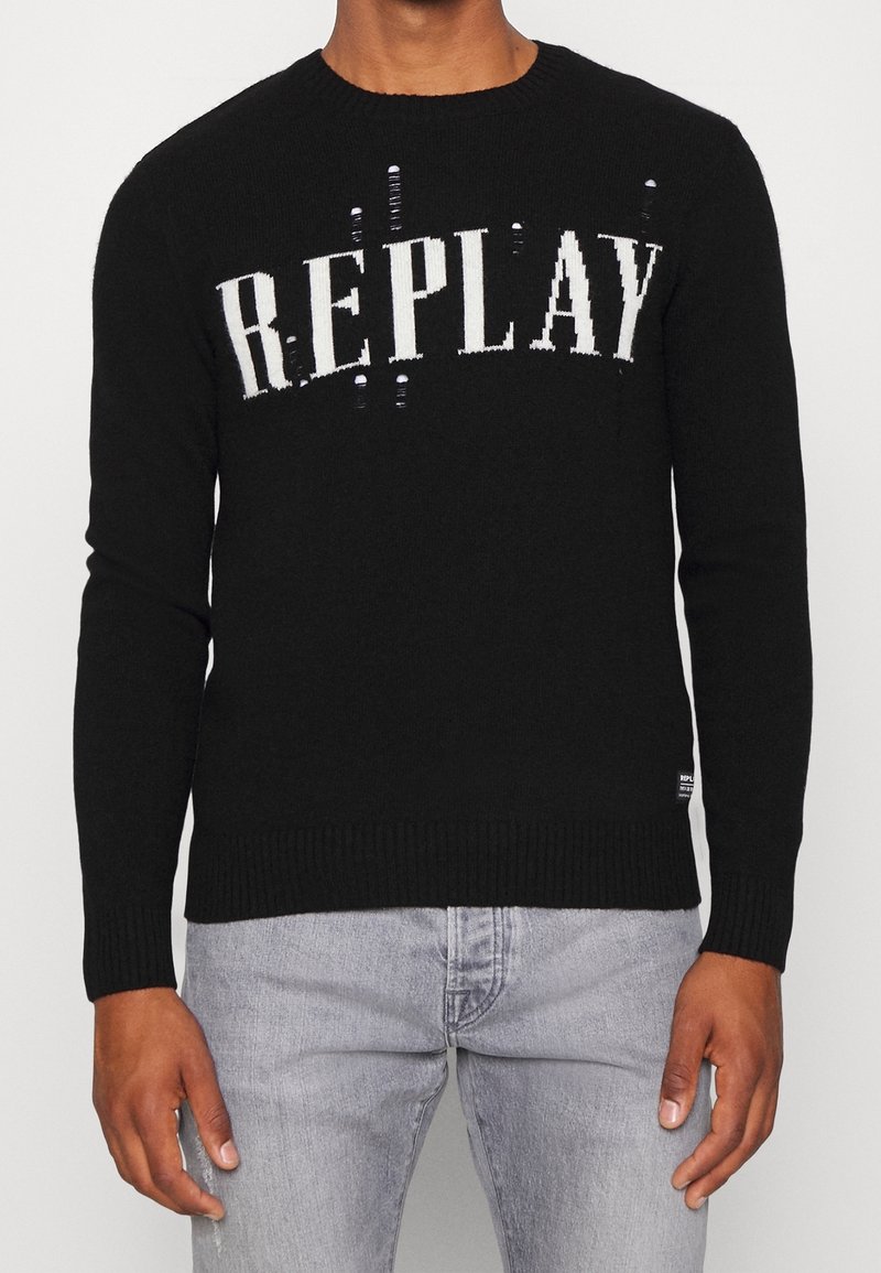 Homme portant un pull noir avec le texte blanc "REPLAY" et un jean gris clair, debout devant un fond uni.