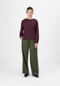 Woman standing in green wide-leg trousers, dark purple long-sleeve blouse, and black shoes against a plain white background.