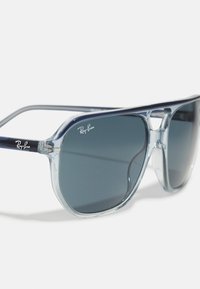 Ochelari de soare cu ramă din acetat transparent și lentile gri închis, având un design rotund și logo-ul argintiu Ray-Ban pe lentila din stânga sus.