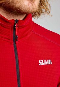 Homme portant une veste zippée rouge avec un motif texturé et un logo blanc "SLINN" sur la poitrine, avec des cheveux blonds bouclés visibles.