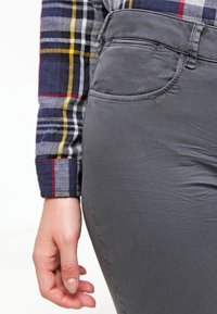 Pantalones elásticos grises con una textura suave y un brillo sutil, que cuentan con un bolsillo frontal y un diseño ajustado, combinados con una camisa de cuadros multicolor.