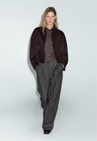 Sort cropped cardigan over en mørk skjorte med knap, parret med grå stribede brede bukser. Glat stof, enkelt design, neutrale farver.
