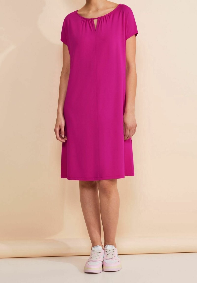 Kvinna klädd i en knälång magentafärgad klänning med korta ärmar och vit-rosa sneakers, stående mot en beige bakgrund.
