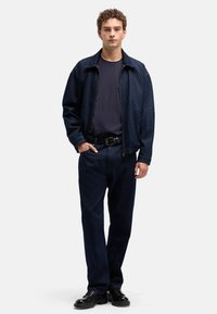 Ciemnoniebieska kurtka dżinsowa z zamkiem na przodzie, w połączeniu z ciemną koszulą i dopasowanymi spodniami. Strój uzupełnia pasek i czarne buty.