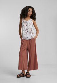 Haut blanc sans manches à motif floral avec des plis, associé à un pantalon ample couleur rouille. Sandales noires avec des détails en liège.