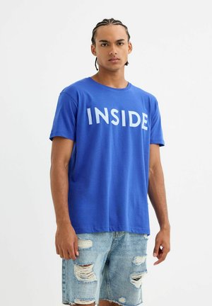 Camiseta de algodón azul con mangas cortas que presenta la palabra "INSIDE" en letras blancas en negrita a lo largo del pecho. Jeans con detalles desgastados.