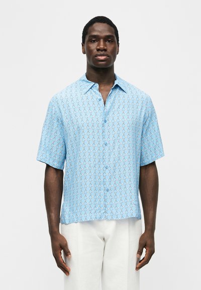 Άνδρας που φοράει γαλάζιο πουκάμισο με κοντά μανίκια και κουμπιά, με κυκλικό σχέδιο και άσπρο παντελόνι, στέκεται μπροστά σε απλό φόντο.