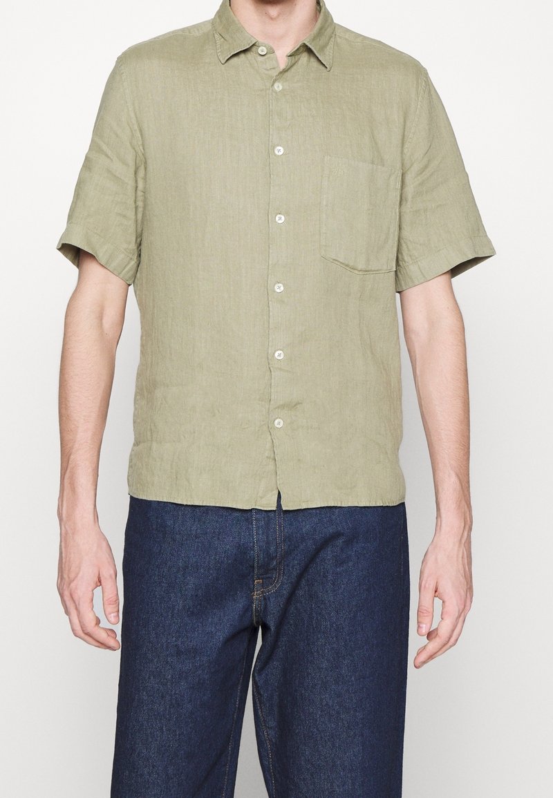 Homme portant une chemise boutonnée à manches courtes vert clair avec poche poitrine et un pantalon en jean bleu foncé, debout devant un fond blanc uni.