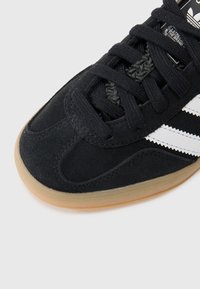 Sort suede sportsko med hvid tre-stribe-design, snørelukning og gummisål. Tekstureret overdel med buede syninger.