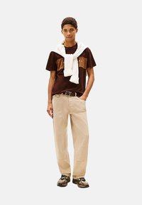 Camiseta marrón con un diseño a bloques de color, suéter crema atado alrededor del cuello, pantalones beige de pierna ancha y zapatillas negras y marrones.