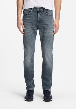 Mand iført falmede blå jeans, hvid t-shirt og marineblå sneakers, stående mod hvid baggrund.