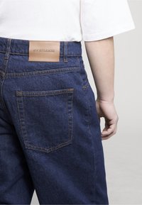 Osoba nosící modré džínové kalhoty s koženou nášivkou a bílým krátkým rukávem, ukazující zadní kapsu a ruku uvolněnou podél těla.