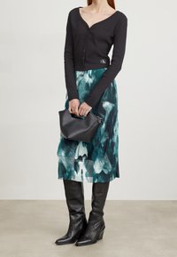 Svart långärmad cardigan, mönstrad knälång kjol i teal och vitt, svarta kilklackstövlar och en liten svart handväska med handtag.