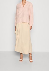 Lichtroze linnen blouse met een V-hals en lange mouwen, gecombineerd met een vloeiende beige maxirok, aangevuld met witte enkelsandalen met riempjes.