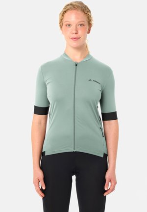 Donna che indossa una maglia da ciclismo verde chiaro a maniche corte con cerniera e pantaloncini da ciclismo neri, in piedi davanti a uno sfondo semplice.