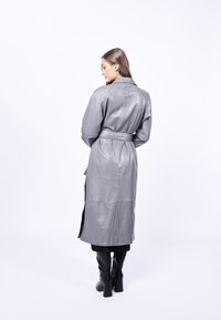 Cappotto in pelle grigia con maniche lunghe, vita con cintura e una spacco sul retro. Texture liscia e design minimalista, con una ventola laterale e colletto.