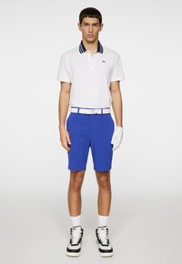 Hvid polo-shirt med marineblå stribet krave, blå shorts, hvidt bælte og sort-hvide træningssko. Inkluderer en hvid golfhandske.