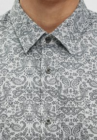 Camisa gris con patrón de cachemira y cuello abotonado, que presenta diseños florales intrincados y una textura suave. Botones negros realzan la parte delantera.