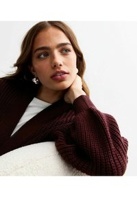 Burgunderfärgad stickad cardigan med en strukturerad mönster, öppen fram, och vida, flödande ärmar. Bärs över en vit topp.