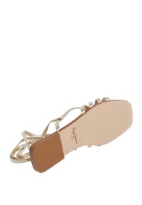Sandalias planas con tiras y un upper de color dorado metálico, planta beige suave y suela marrón texturizada. El diseño notable incluye detalles decorativos en forma de lazo.
