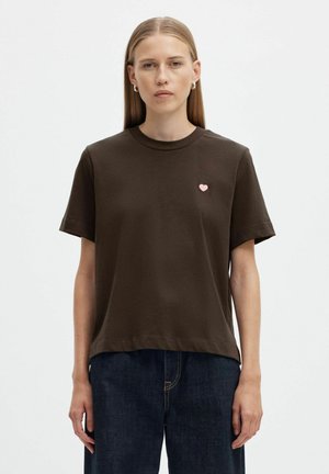 Femme aux cheveux raides portant un t-shirt marron foncé à manches courtes avec un petit cœur rose sur la poitrine et un jean en denim foncé.