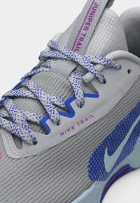 Κοντινή λήψη ενός γκρι παπουτσιού Nike Trail για τρέξιμο, με κορδόνια μπλε με στίγματα και μωβ κείμενο στη γλώσσα και λεπτομέρειες στο πλάι με σχέδιο διαδρομής.
